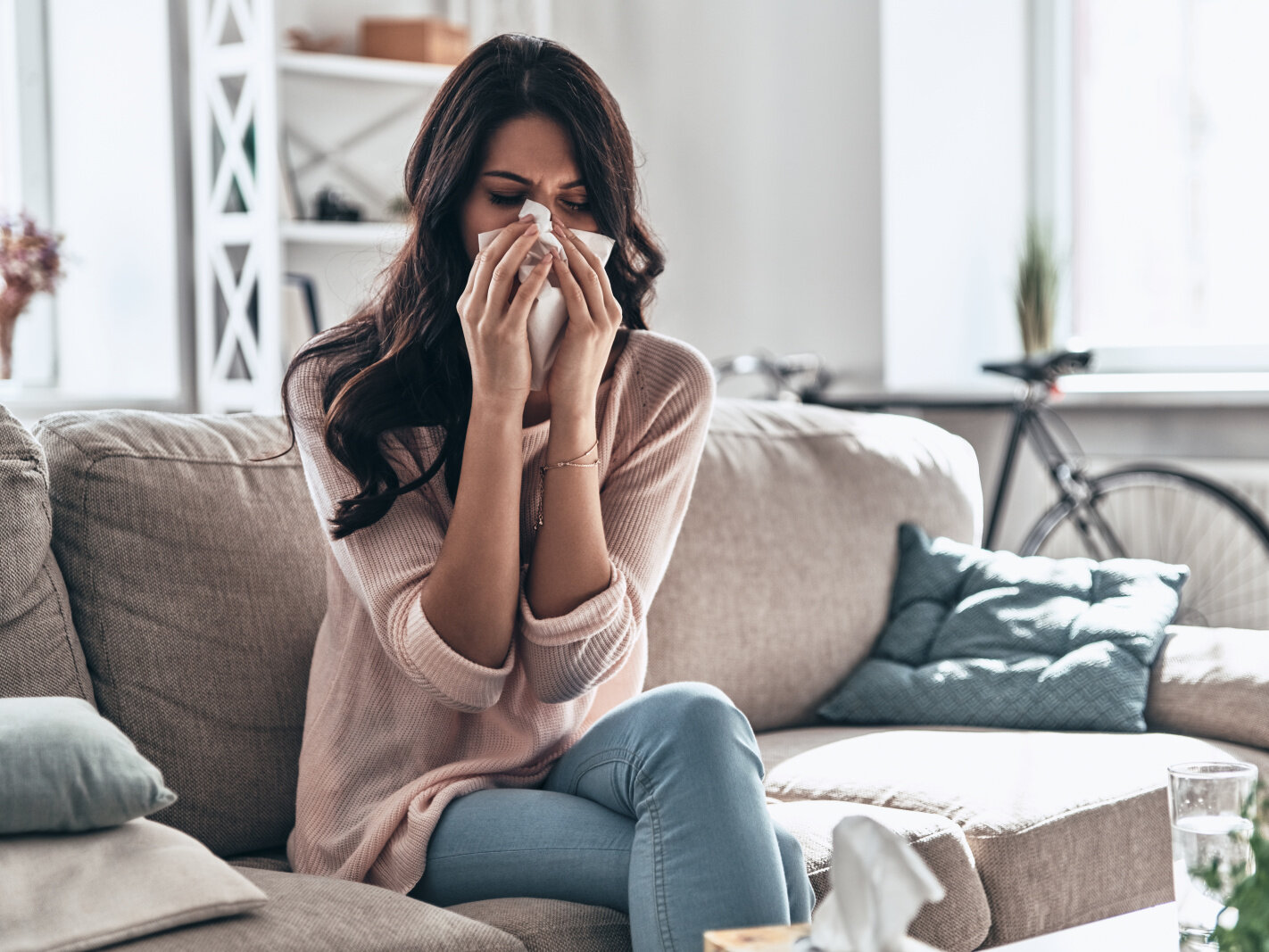 AdobeStock_233210436 Cold and flu. Sick young woman blowing the nose using tissue paper while sitting on the sofa at home, Woman at home with flu or allergy symptoms cleaning her nose